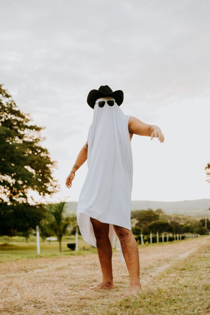 Man in a cowboy hat and ghost costume posing on a rural road with a comedic touch.